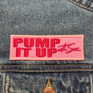 Könnte beinhalten: Eine rosafarbene Jeansjacke mit einem rosafarbenen Patch, auf dem in roten Buchstaben "PUMP IT UP with Sue" steht.