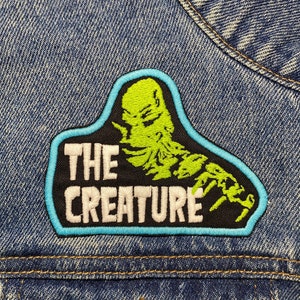 Könnte beinhalten: Eine blaue Jeansjacke mit einem schwarzen und grünen gestickten Aufnäher, auf dem "THE CREATURE" in weißer Schrift steht.