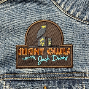 Pode incluir: Um remendo bordado marrom e azul com o texto "Night Owls with Jack DeJohnette" em uma jaqueta de jeans.