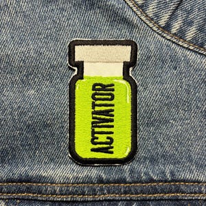 May include: Embroidered patch featuring a vial design with the word "ACTIVATOR" in black capital letters on a bright green background. The patch has a black border and a white top, set against a denim jacket.