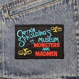 May include: A black and red embroidered patch with the text "Captain Spaulding's Museum of Monsters and Madmen" on a blue denim background.