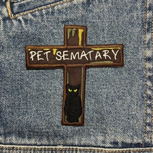 Puede incluir: Una cruz de madera marrón con el texto "Pet Sematary" en ella. Un gato negro con ojos amarillos está detrás de la cruz.
