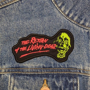 Puede incluir: Un parche bordado negro y verde con el texto "The Return of the Living Dead" en rosa y una cara de zombie verde.