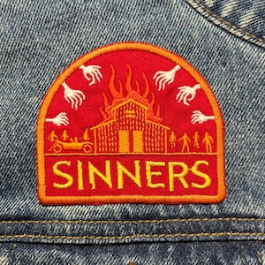 May include: A red and orange embroidered patch with the word "SINNERS" in yellow. The patch features a burning building, white hands, and a vintage car. The patch is sewn onto a blue denim jacket.