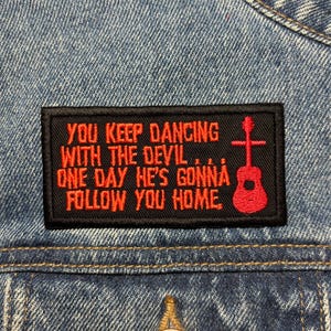 Peut inclure: Un écusson rectangulaire noir avec du texte rouge et un dessin de guitare. Le texte dit : "YOU KEEP DANCING WITH THE DEVIL... ONE DAY HE'S GONNA FOLLOW YOU HOME." L'écusson est cousu sur une veste en jean.
