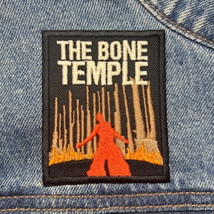 Könnte beinhalten: Gestickter Aufnäher mit dem Text "THE BONE TEMPLE" über einer orangefarbenen Figur vor einer Waldszene mit Flammen. Der Aufnäher ist rechteckig mit einem schwarzen Rand und auf eine Jeansjacke genäht.