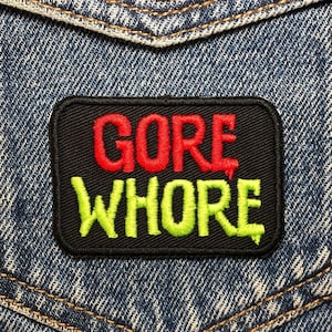 Peut inclure: Une veste en jean avec un patch noir qui dit "GORE WHORE" en lettres rouges et vertes.
