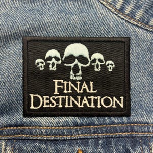Może przedstawiać: Czarna prostokątna naszywka z białym haftowanym wzorem trzech czaszek i napisem "FINAL DESTINATION". Naszywka jest przyszyta do niebieskiej kurtki dżinsowej.