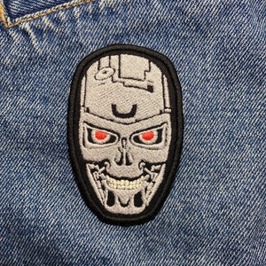 Puede incluir: Un parche bordado en blanco y negro de una calavera de robot con ojos rojos. El parche está sobre un fondo de mezclilla azul.