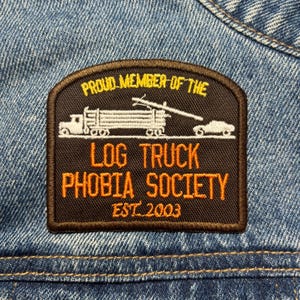 Puede incluir: Un parche rectangular con borde marrón y centro negro, con el texto "PROUD MEMBER OF THE LOG TRUCK PHOBIA SOCIETY EST. 2003". El parche incluye una ilustración blanca de un camión maderero y un coche. El parche está cosido sobre una tela vaquera azul.
