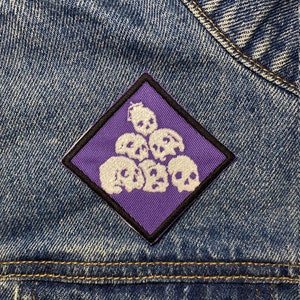 May include: Purple diamond-shaped patch with white embroidered skulls. The patch is sewn onto a blue denim jacket.