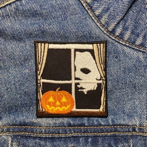 Op de afbeelding: Een bruin en zwart geborduurd patch met een vensterframe, een jack-o'-lantern en een witte geest die door het venster kijkt.