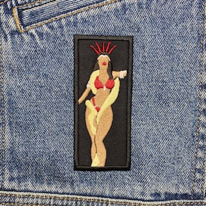 May include: A black and white embroidered patch of a woman wearing a red bikini and a yellow sash. She has a crown of red stars on her head. The patch is on a blue denim background.
