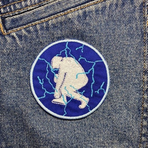 Könnte beinhalten: Ein blauer und weißer gestickter Aufnäher mit einer weißen Silhouette einer Person, die in einem Blitz-Muster steht.