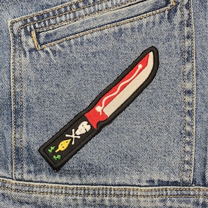 May include: A black and red embroidered patch of a knife with a skull and crossbones on a blue denim jacket.