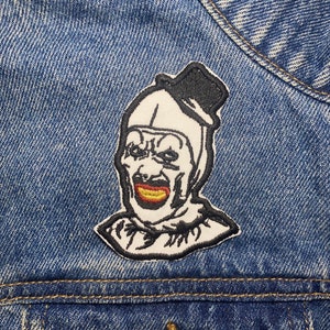 May include: A white embroidered patch of a creepy clown with a black top hat, a yellow smile, and black eyes. The patch is sewn onto a blue denim jacket.