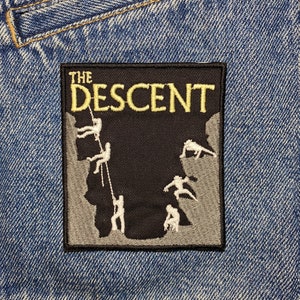 Op de afbeelding: Zwart-wit geborduurde patch met de tekst "THE DESCENT" in gele letters. De patch toont een groep mensen die aan een klif abseilen.