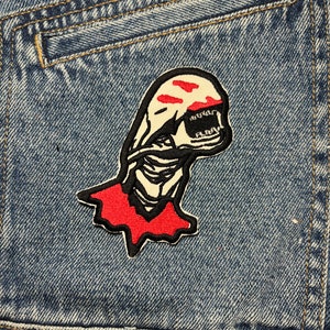 May include: A black and white embroidered patch of a creature with red markings on a blue denim jacket. The creature has a long, thin body and a large head with a mouth full of teeth. The patch is sewn onto the pocket of the jacket.