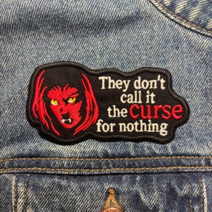 May include: Embroidered patch with a black background and the text "They don't call it the CURSE for nothing." The patch features a red devil face with yellow eyes and red hair. The patch is sewn onto a denim jacket.