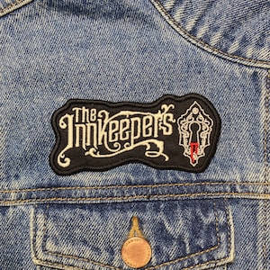 Può includere: Una toppa ricamata in bianco e nero con il testo "The Innkeepers" in un font stilizzato. La toppa presenta un buco della serratura con una goccia di sangue rosso.