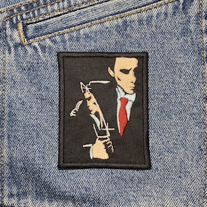 Peut inclure: Patch brodé noir et blanc d'un homme en costume avec une cravate rouge, tenant une cigarette dans sa main droite. Le visage de l'homme est partiellement masqué par la cigarette.