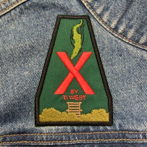 May include: A green, triangular patch with a red X over a yellow flame. The text "BY THE WEST" is below the X.