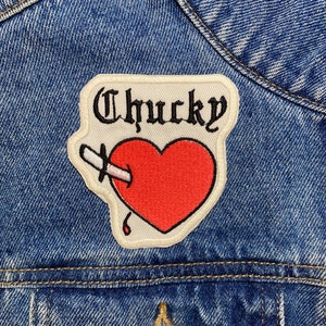 Può includere: Una toppa bianca con la parola "Chucky" in lettere nere e un cuore rosso con un coltello che lo attraversa. La toppa è cucita su una giacca di jeans blu.