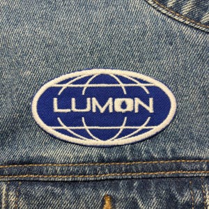 May include: An embroidered patch with the word "LUMON" in white capital letters. The patch is oval and features a white globe design on a blue background. It is sewn onto a denim jacket.