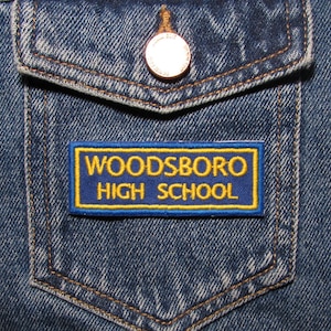 Puede incluir: Primer plano de un bolsillo de chaqueta vaquera azul con un parche rectangular. El parche, con fondo azul, muestra texto bordado en amarillo: "WOODSBORO HIGH SCHOOL". La chaqueta tiene un botón cobrizo y costuras.
