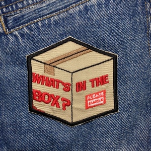 May include: A brown cardboard box with the text "What's in the box?" in red letters. A red rectangle with the text "Please handle with care" is on the side of the box. The box is sewn onto a blue denim fabric.