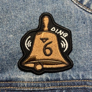 Può includere: Toppa a forma di campana marrone con il numero 6 all'interno, circondata da un bordo nero. La parola "DING" è arcuata sopra. La toppa è cucita su una giacca di jeans.