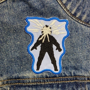 Op de afbeelding: Een witte en blauwe geborduurde patch met een zwart silhouet van een persoon met witte lichtstralen die uit hun hoofd komen.