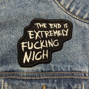 May include: A black embroidered patch with the text "THE END IS EXTREMELY FUCKING NIGH" in white lettering. The patch is sewn onto a blue denim jacket, with visible stitching and fabric texture.
