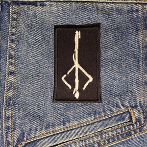 Peut inclure: Un écusson brodé noir et blanc avec un symbole stylisé d'une tête de chèvre avec des cornes et un pentagramme. L'écusson est cousu sur une veste en jean bleue.