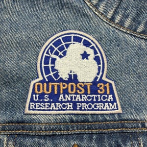 Może przedstawiać: Naszywka haftowana w kolorze niebieskim i białym z tekstem "Outpost 31 U.S. Antarctica Research Program". Naszywka przedstawia stylizowane zdjęcie Antarktydy w kółku.