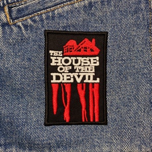 Może przedstawiać: Czarno-czerwona naszywka haftowana z tekstem "The House of the Devil" i czerwoną sylwetką domu z kominem. Naszywka znajduje się na niebieskim denimie.
