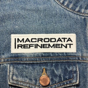 Puede incluir: Un parche blanco cosido en una chaqueta vaquera azul con el texto "MACRODADA REFINEMENT" en negro.