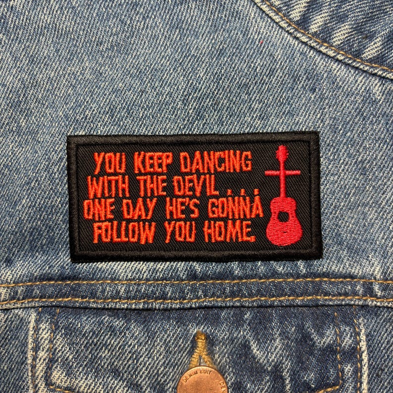 K&ouml;nnte beinhalten: Ein schwarzer rechteckiger Aufn&auml;her mit rotem Text und einer Gitarren-Grafik. Der Text lautet: "YOU KEEP DANCING WITH THE DEVIL... ONE DAY HE'S GONNA FOLLOW YOU HOME." Der Aufn&auml;her ist auf eine Jeansjacke gen&auml;ht.