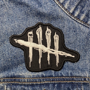 Op de afbeelding: Een geborduurde patch in zwart en wit met het iconische 'Dead by Daylight'-logo. Het logo bestaat uit vijf witte verticale lijnen met een witte schuine streep bovenaan. De patch is genaaid op een blauw denim jack.