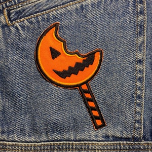Könnte beinhalten: Ein Bügeleisen-Aufnäher mit Halloween-Motiv, der eine angebissene orangefarbene Kürbis-Lollipop mit einem schwarzen Kürbisgesicht auf einem blauen Jeans-Hintergrund zeigt.