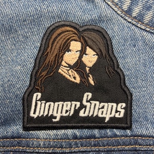 Puede incluir: Parche bordado con dos figuras femeninas estilizadas con cabello largo, en tonos marrón y negro, sobre un fondo negro. El parche dice "Ginger Snaps" en letras blancas. El parche está cosido a una chaqueta vaquera.