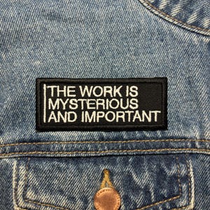 May include: Black and white embroidered patch with the text "The work is mysterious and important" on a blue denim jacket.