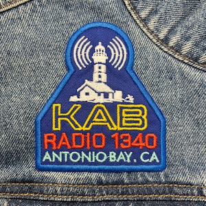 Pode incluir: Um patch azul, branco, amarelo e vermelho com um desenho de farol com o texto "KAB RADIO 1340 ANTONIO BAY, CA". O patch é costurado em uma jaqueta jeans.
