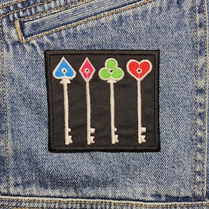 Może przedstawiać: Czarna, haftowana naszywka z czterema kluczami z kolorowymi wierzchołkami: niebieskim, różowym, zielonym i czerwonym. Klucze są umieszczone na czarnym tle i przyszyte do dżinsowej kurtki.