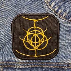 May include: A black patch with a yellow embroidered symbol. The symbol is a circle with a line through the centre and a smaller circle inside the larger circle.