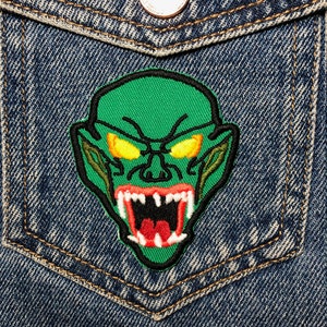 Può includere: Toppa ricamata con la faccia di un mostro verde con occhi gialli e una grande bocca aperta con denti affilati. La toppa è cucita su una giacca di jeans blu.