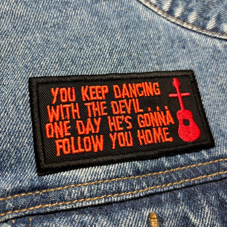 K&ouml;nnte beinhalten: Ein schwarzer rechteckiger Aufn&auml;her mit rotem gesticktem Text und einer Gitarren-Grafik. Der Text lautet: "YOU KEEP DANCING WITH THE DEVIL... ONE DAY HE'S GONNA FOLLOW YOU HOME." Der Aufn&auml;her ist auf eine Jeansjacke gen&auml;ht.