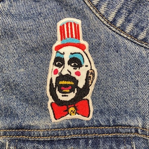 Könnte beinhalten: Ein gestickter Aufnäher mit einem Clown mit Zylinder, Bart und roter Fliege. Der Clown hat ein weißes Gesicht mit schwarzen Augen und einer rosa Nase, und der Hut ist mit roten, weißen und blauen Streifen verziert.