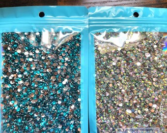 May include: Two bags of clear plastic filled with small, round, iridescent rhinestones. One bag contains teal rhinestones, and the other bag contains clear rhinestones.