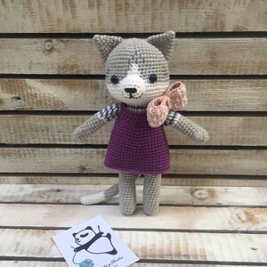 Könnte beinhalten: Eine gehäkelte graue Katze, die ein lila Kleid und eine rosa Schleife trägt. Die Katze steht auf einer Holzoberfläche. Das Bild enthält auch eine kleine Karte mit dem Text "HelenToy Studio Handmade Toys".
